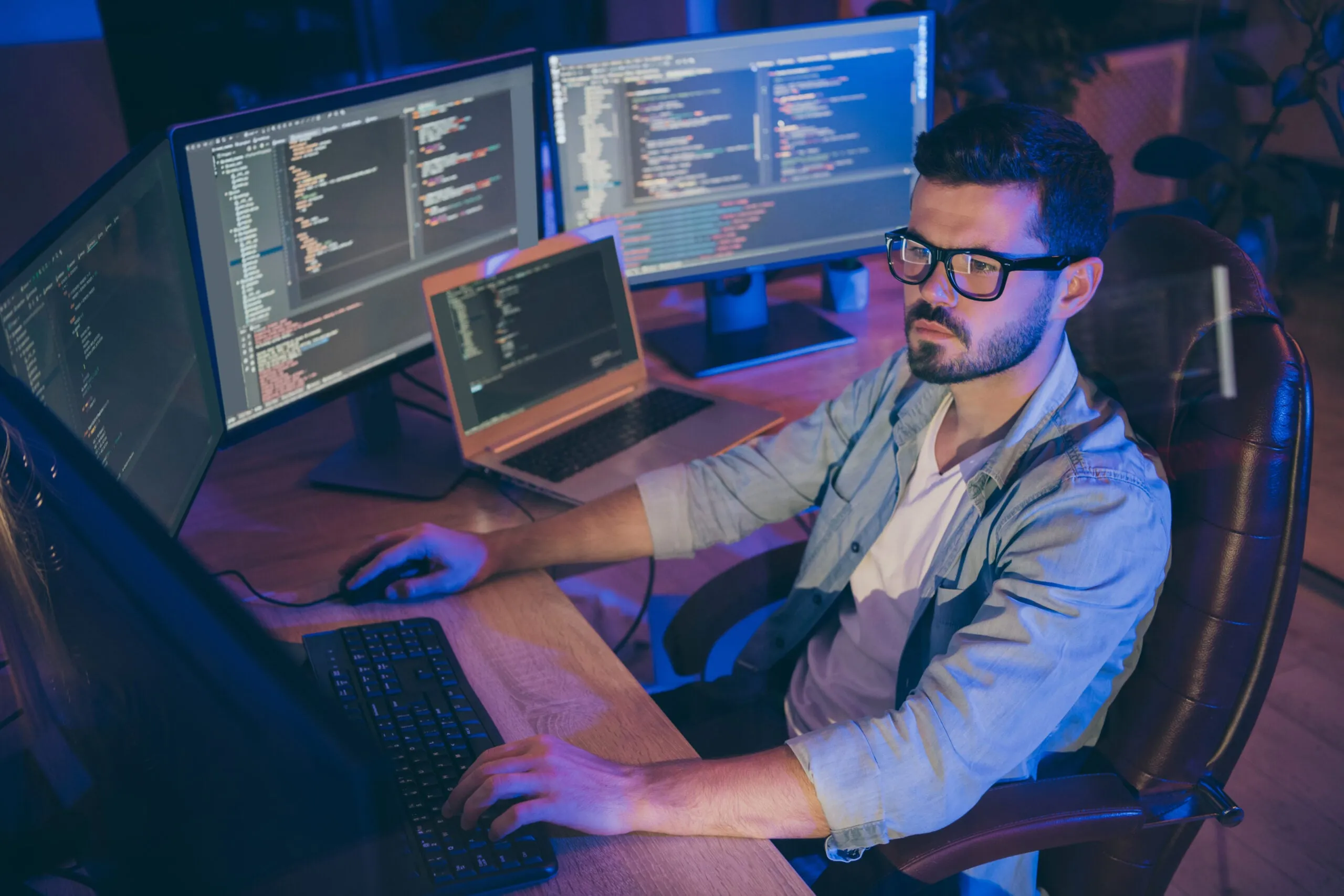 A focused professional in glasses working at a desk with two monitors displaying code alongside an open laptop, lit by warm ambient light.