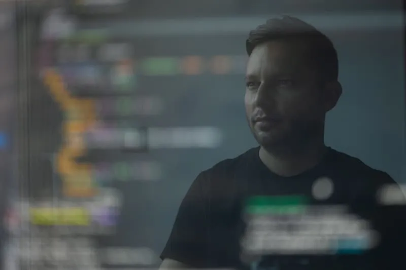 Portrait of a person in a dark shirt looking to the side, with blurred code and data visualizations on a monitor behind them, lit in cool tones.