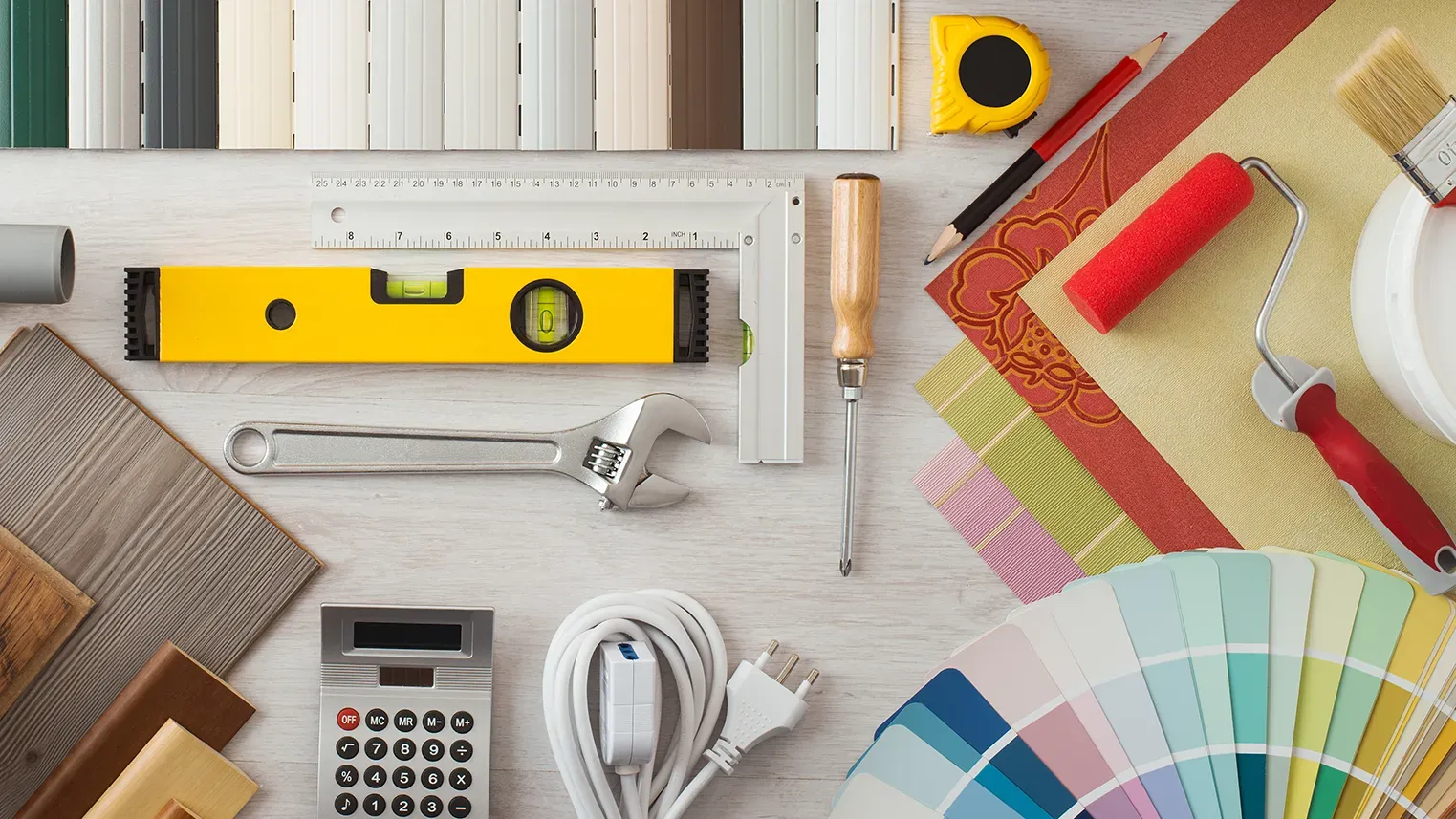 Overhead flat-lay of handyman tools and supplies on a light wood surface — spirit level, wrench, paint roller, measuring tape, paint color fan, calculator, and cables.